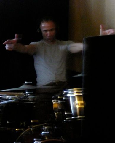 Drummer playing, arms outstretched over kit. He wears a light shirt and headphones in a dark room.