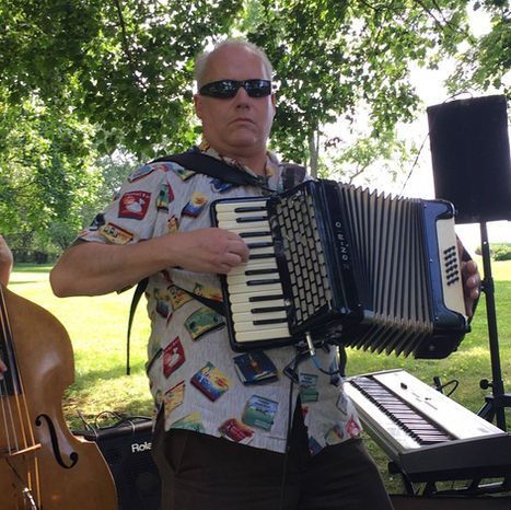 Man playing an accordion outside, wearing sunglasses and a patterned shirt, with a double bass and keyboard nearby.