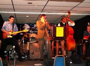 Blues band performing on a stage: Guitarist, trumpet player, bassist, and drummer play music. Low lighting.