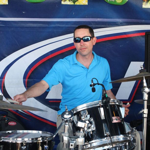 Man in sunglasses plays drums at an outdoor event, wearing a blue shirt.