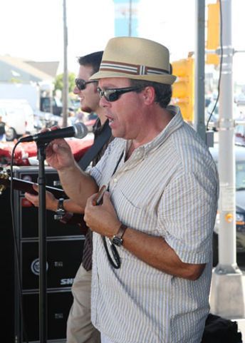 Two men singing outdoors: one in a fedora and sunglasses, the other wearing sunglasses and holding a guitar.