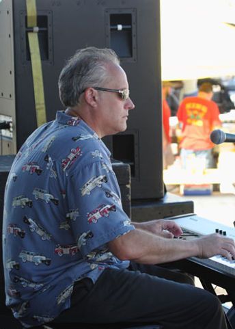Man playing a keyboard outdoors, wearing a blue shirt with car print. He's wearing sunglasses.