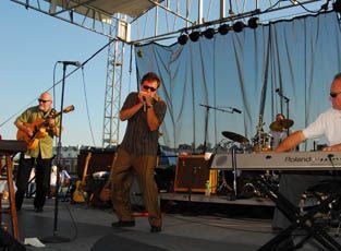 Band performing on outdoor stage: guitarist, harmonica player, keyboardist. Blue sky, concert setting.