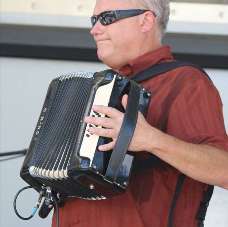 Man playing a black and white accordion, wearing sunglasses and a red shirt. Outdoor setting.