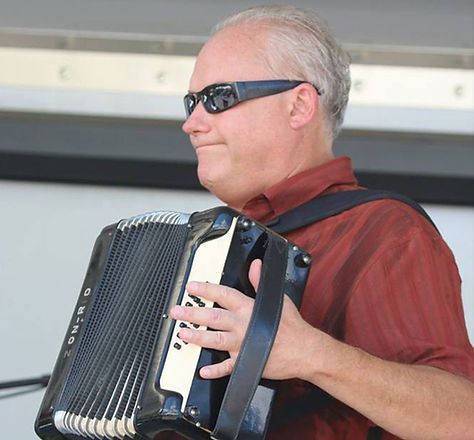 Man in sunglasses playing an accordion outdoors, wearing a red shirt.