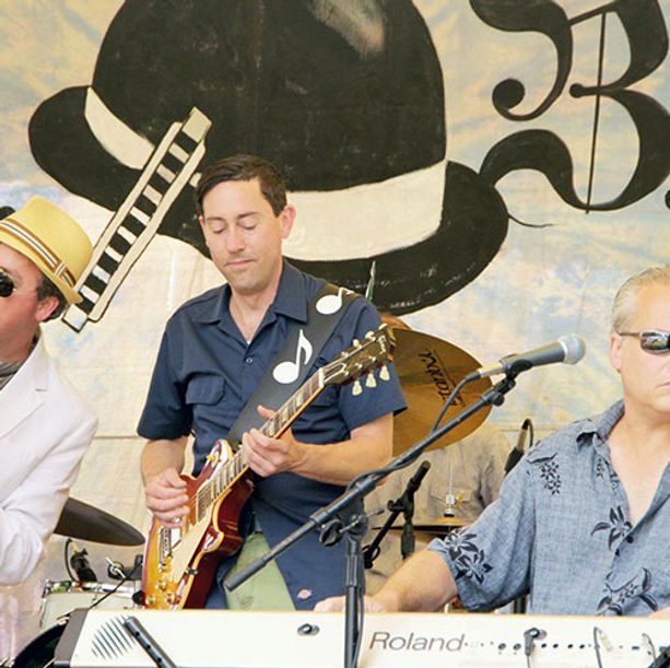 Band performing on stage: guitarist, keyboardist, and drummer play in front of a painted backdrop with a hat.