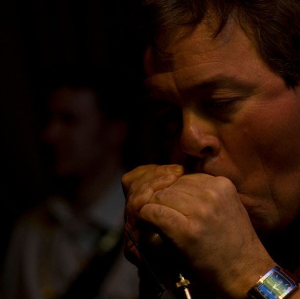 Man playing harmonica, lit by stage light. He's blowing into the instrument, hand close up. Dark background with a figure.