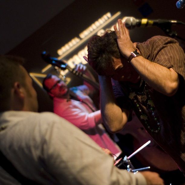 Man on stage with hands on head, bandmates and Drexel University banner in background.