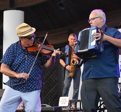 Musicians perform onstage: violin, accordion, and saxophone. Blue and white clothing, park setting.