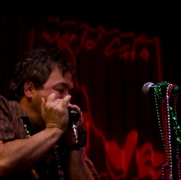 Man playing harmonica onstage at the World Cafe, wearing beads; red lighting.