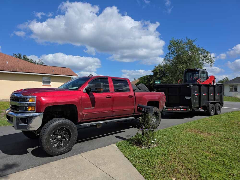 Red pickup truck pulling a trailer with landscaping equipment on a sunny day in a residential area.