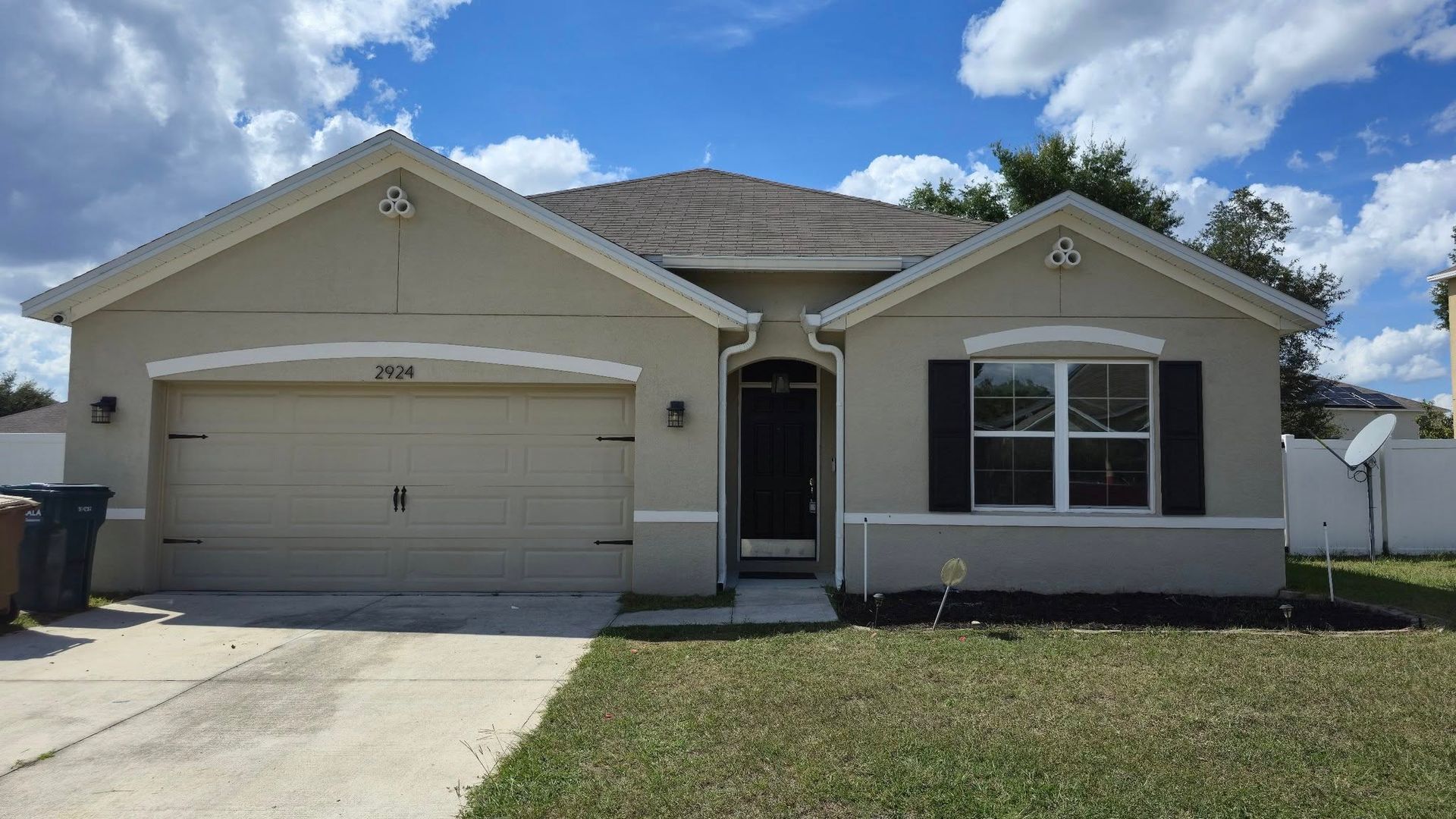 Tan house with a garage, black shutters, and a gray roof under a blue sky with clouds.