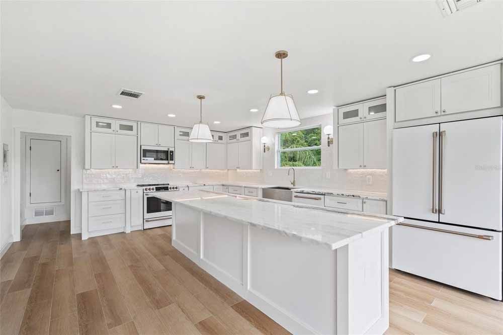 White kitchen with island, cabinets, appliances, and wood flooring. Two pendant lights hang above.