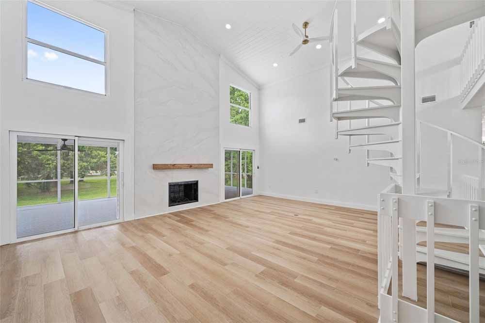 Empty living room with wood floors, fireplace, spiral staircase, and large windows.