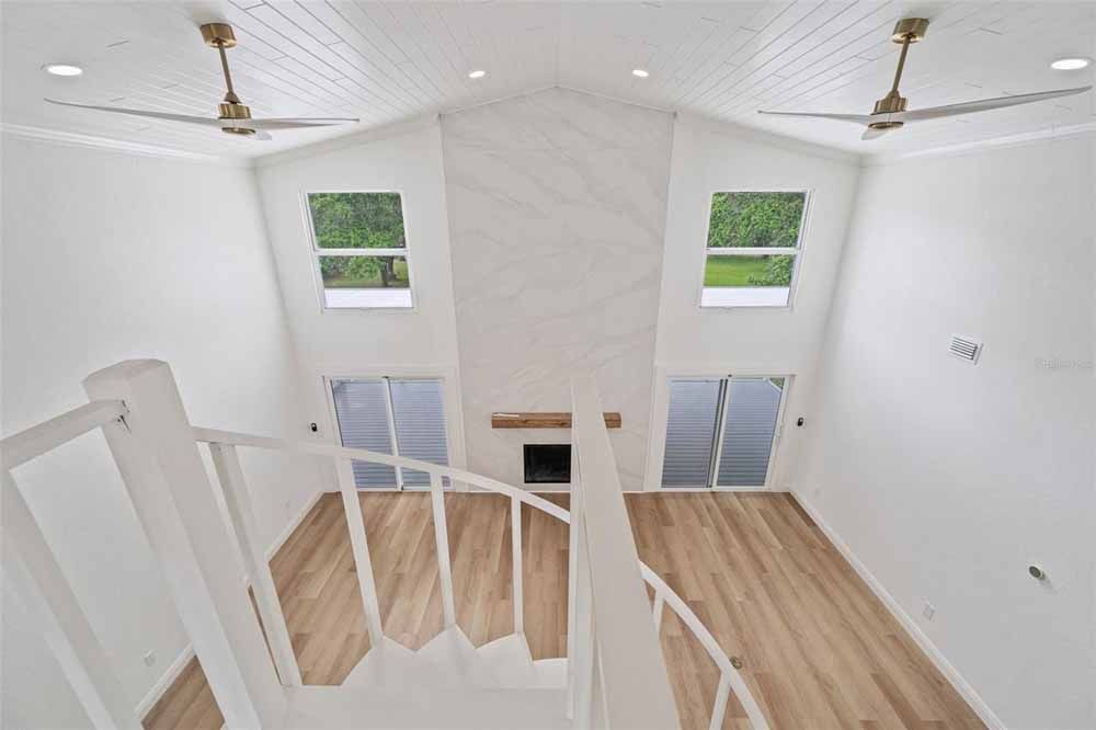High-ceilinged white room with wood floors, two windows, two sliding doors, fireplace, and two ceiling fans.