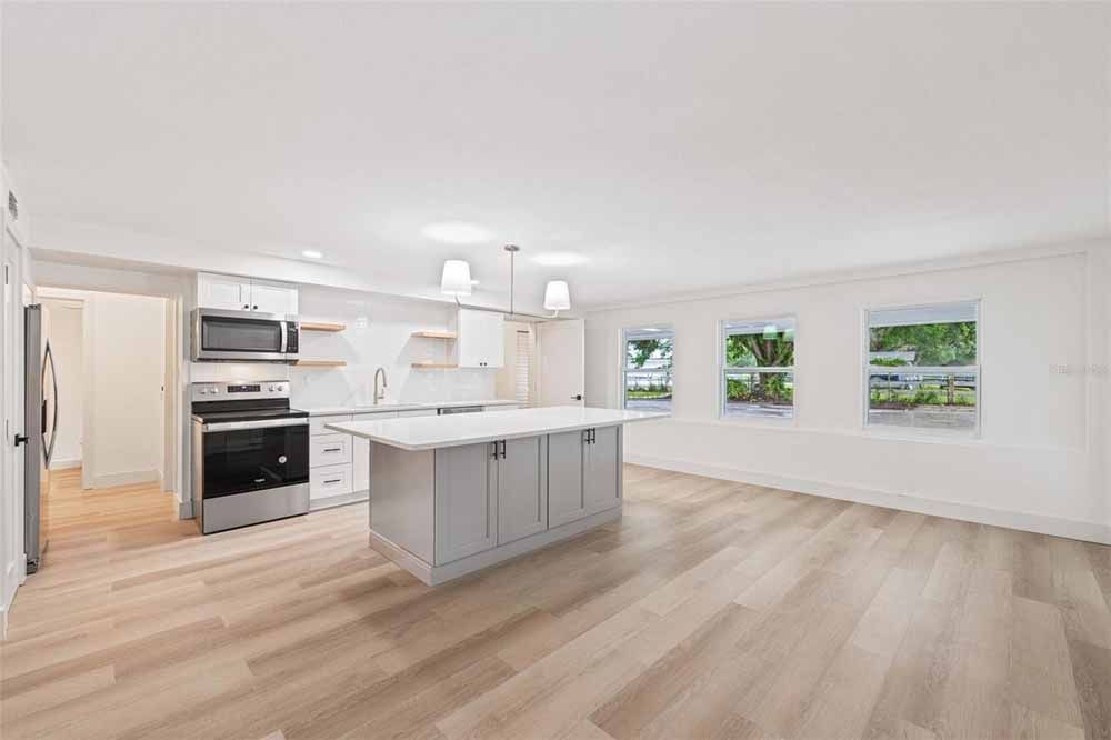 Open-concept kitchen with light wood floors, white cabinets, and a gray island. Three windows let in natural light.