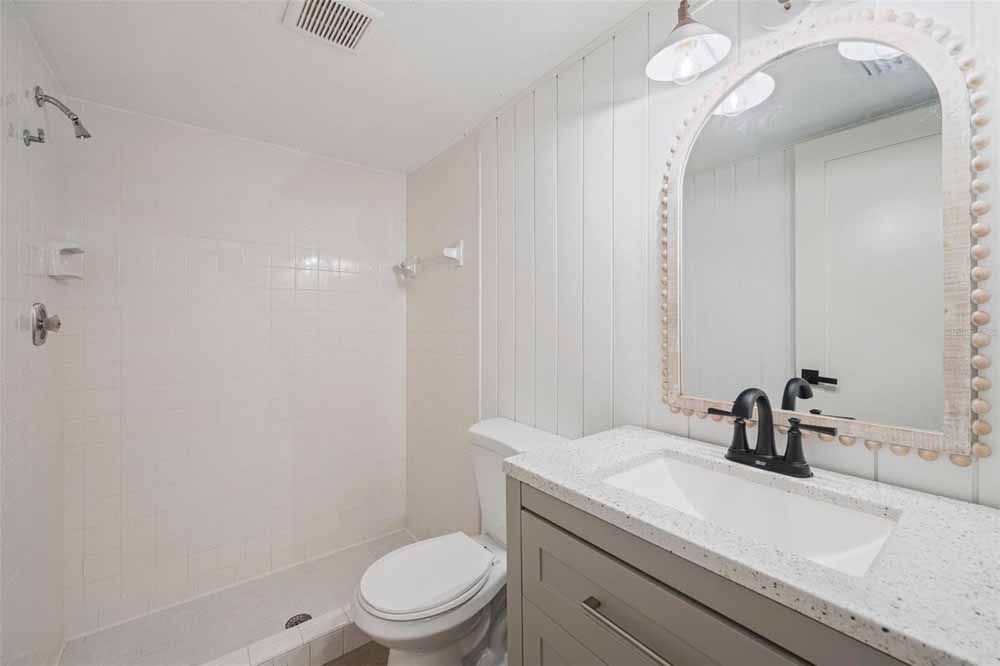White bathroom with a shower, toilet, vanity with mirror, and vent.