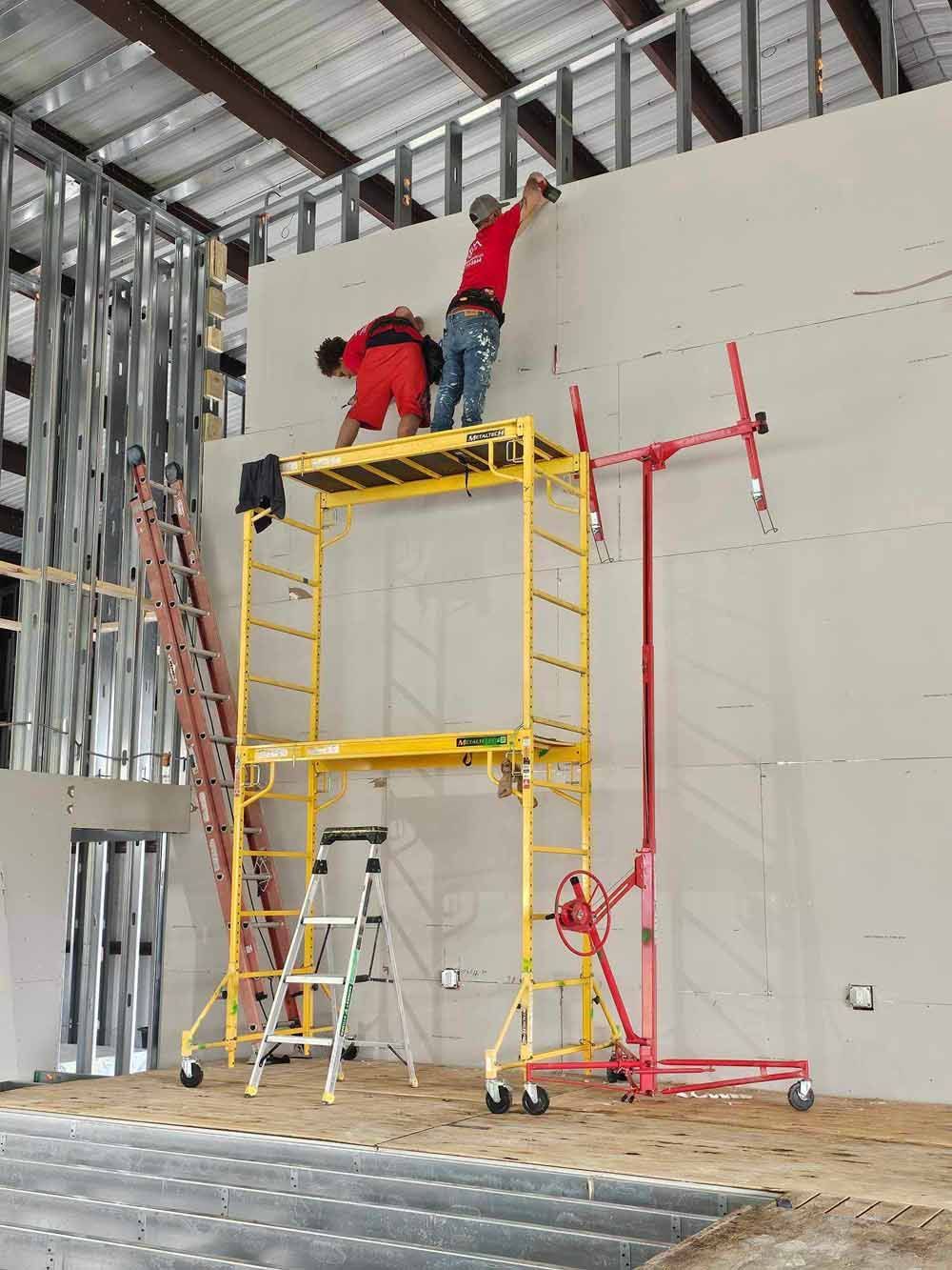 Two workers installing drywall in a large industrial space, using a scaffold and drywall lift.