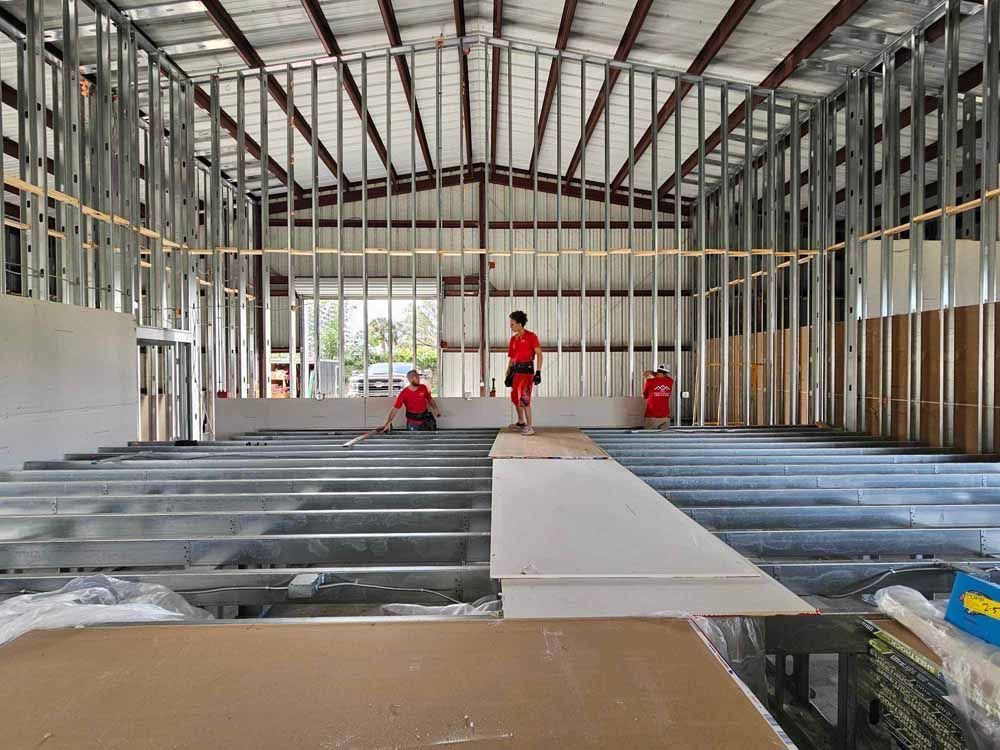 Interior view of a building under construction, workers installing drywall. Steel framing visible.