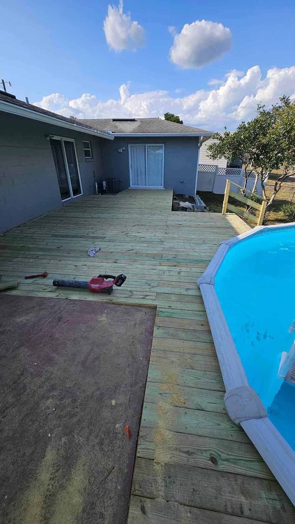 Newly constructed wooden deck next to an above-ground pool and a house with blue sky and clouds.