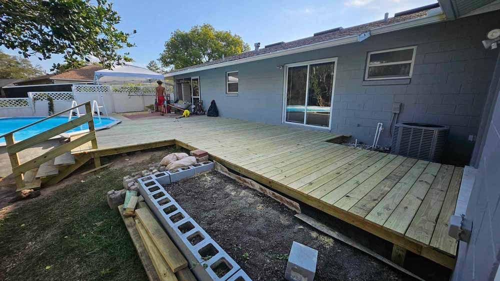 Newly built wooden deck extending from a gray house, with a swimming pool and construction materials visible.