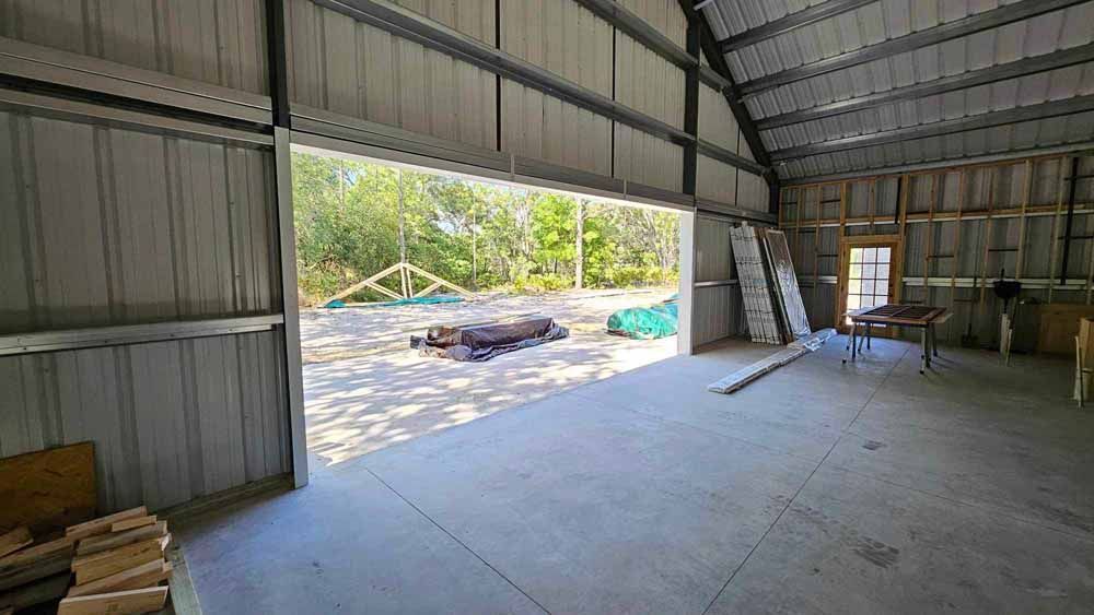 Interior of a metal building with an open door facing a wooded area. Concrete floor, wooden studs on one side.