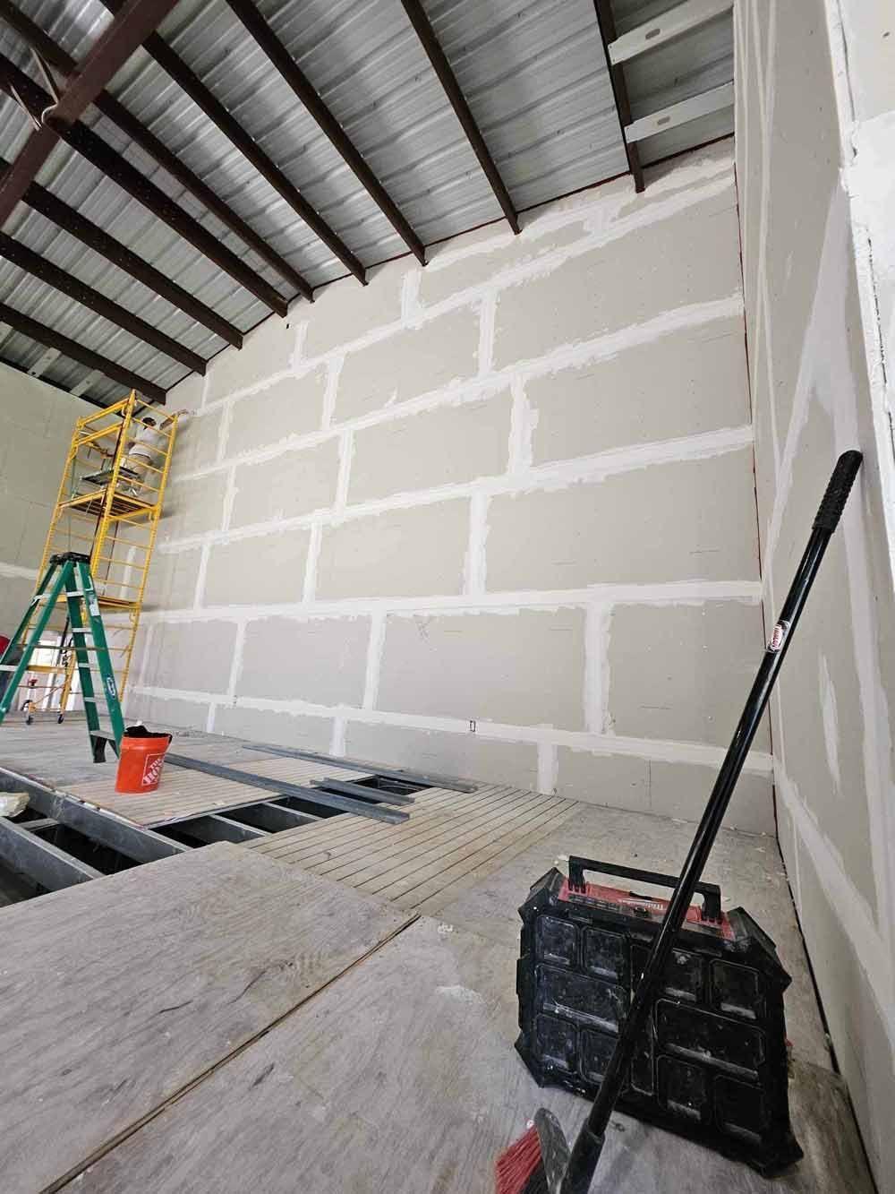 Interior of a building under construction, with drywall installation on a wall, tools and scaffolding present.