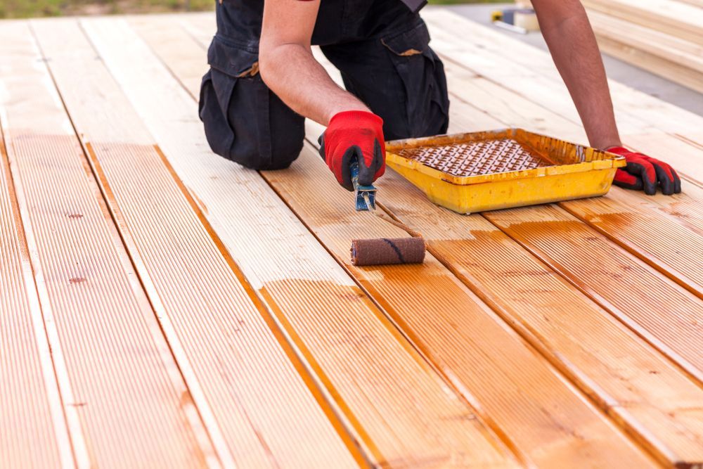 Person applying wood stain to a wooden deck with a roller, wearing gloves.