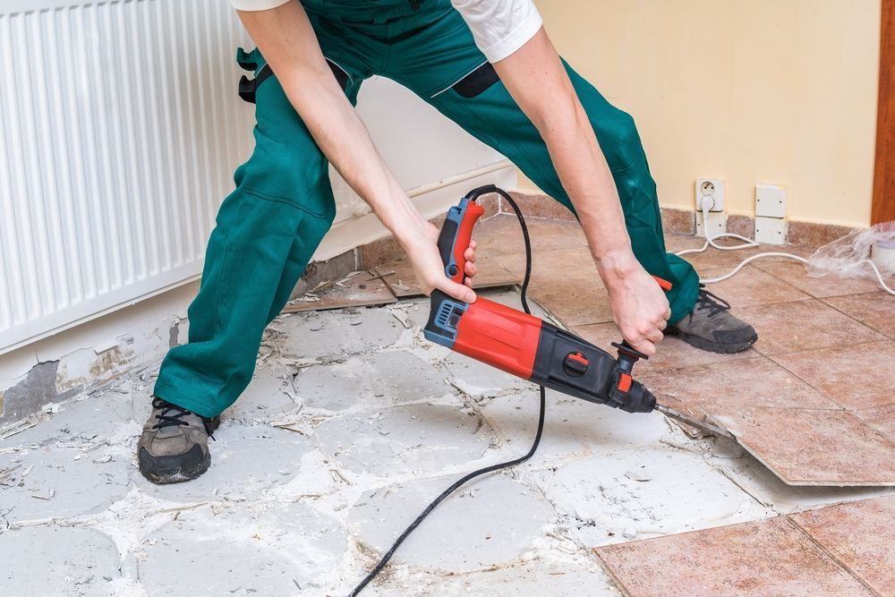Person in green overalls using a red power tool to remove floor tiles in a room.