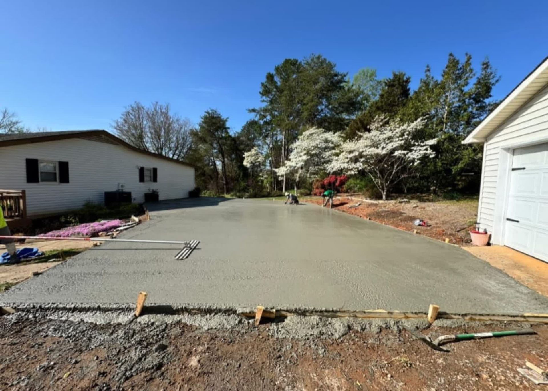 Freshly poured concrete driveway in front of a white house and garage, with trees and blue sky.