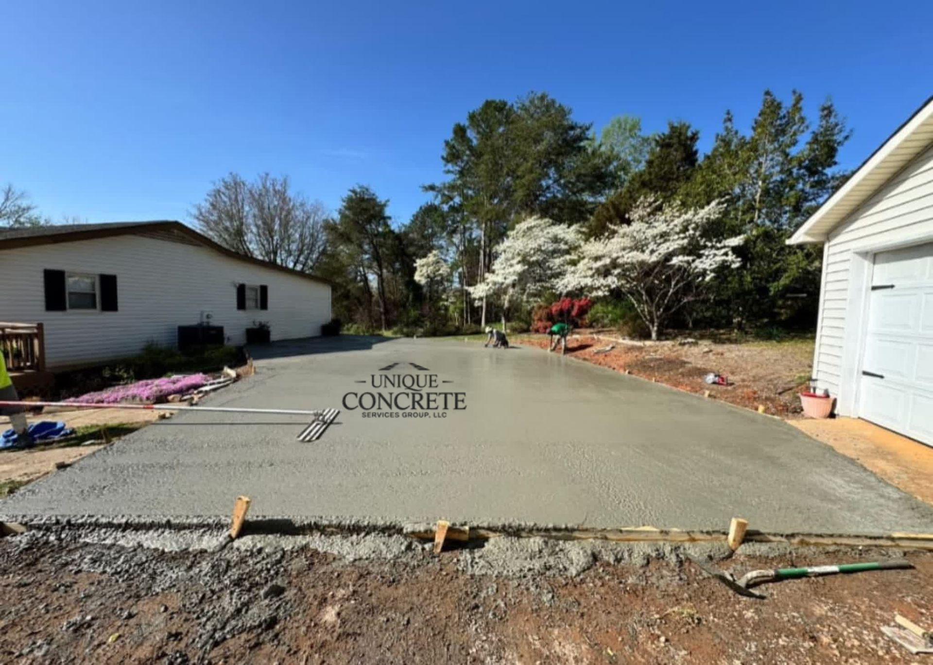 Freshly poured concrete driveway in front of a white house and garage, with trees and blue sky.