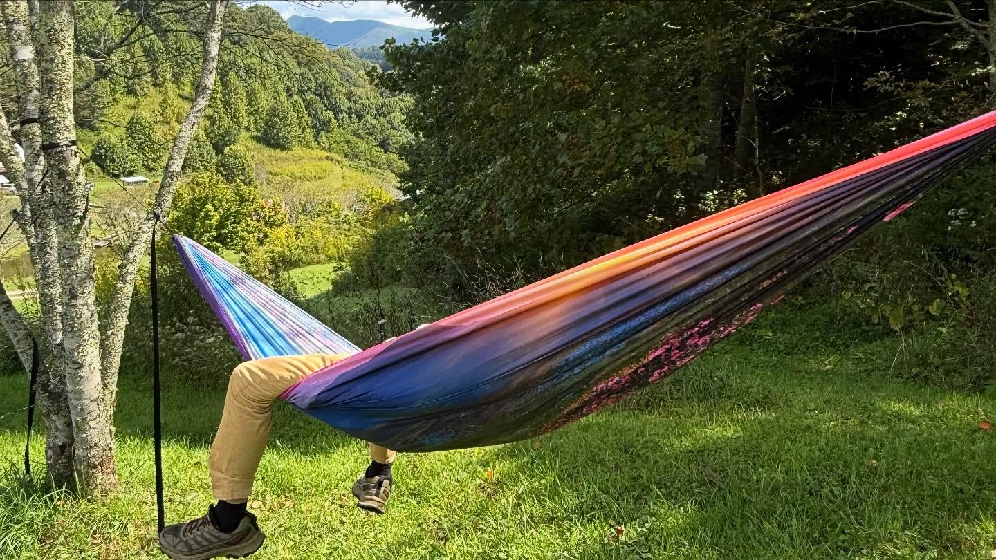 Person relaxing in a colorful hammock strung between trees, set in a green, hilly landscape.
