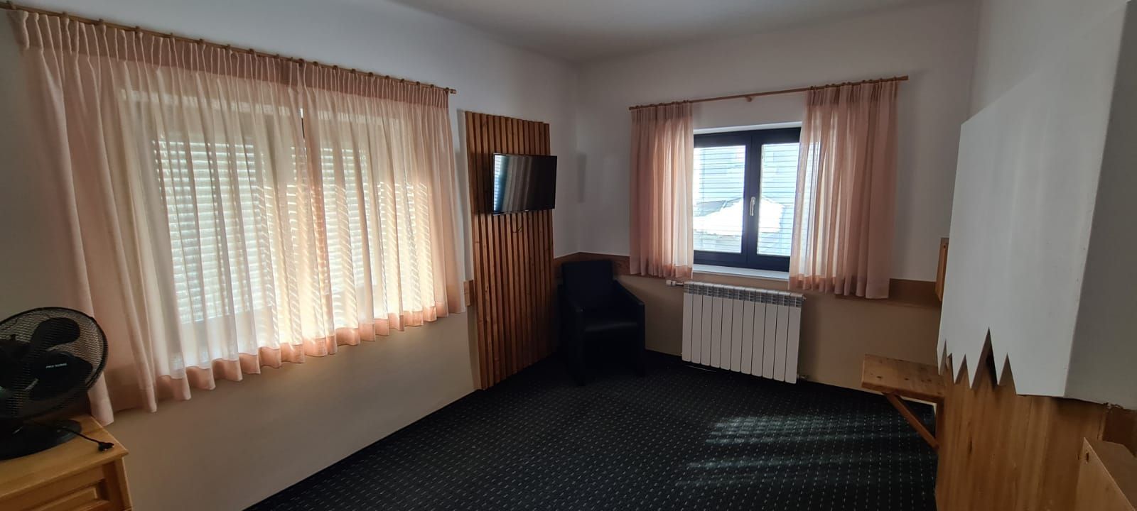 Ein Zimmer mit zwei Fenstern, einem Stuhl, einem Ventilator und einem Fernseher.