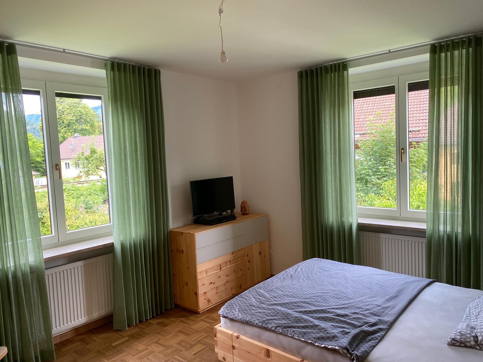 Ein Schlafzimmer mit Bett, Kommode, Fernseher und grünen Vorhängen.