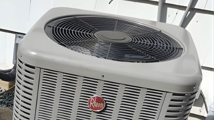 A beige Rheem outdoor air conditioning unit sits on the ground next to the white siding of a house.