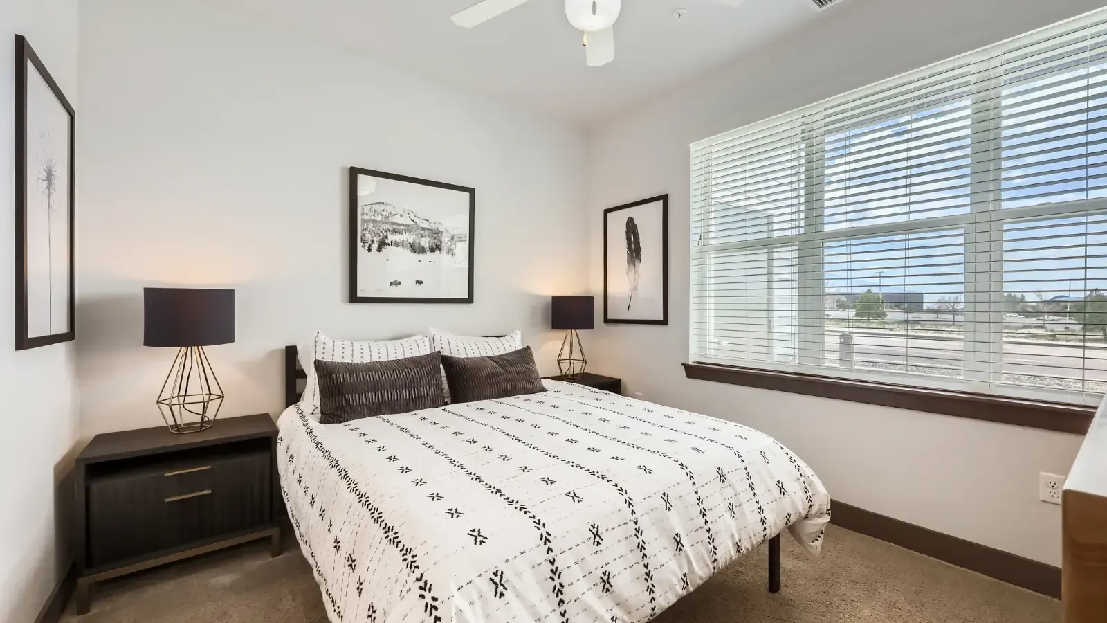 Bedroom with a queen bed, two dark nightstands, two lamps, framed art, and a large window with horizontal blinds.
