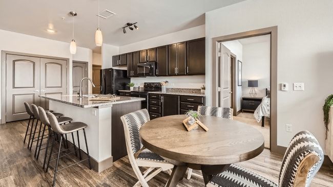 Modern kitchen with island, black cabinets, wood floors, and round dining table. Open doorway to bedroom.