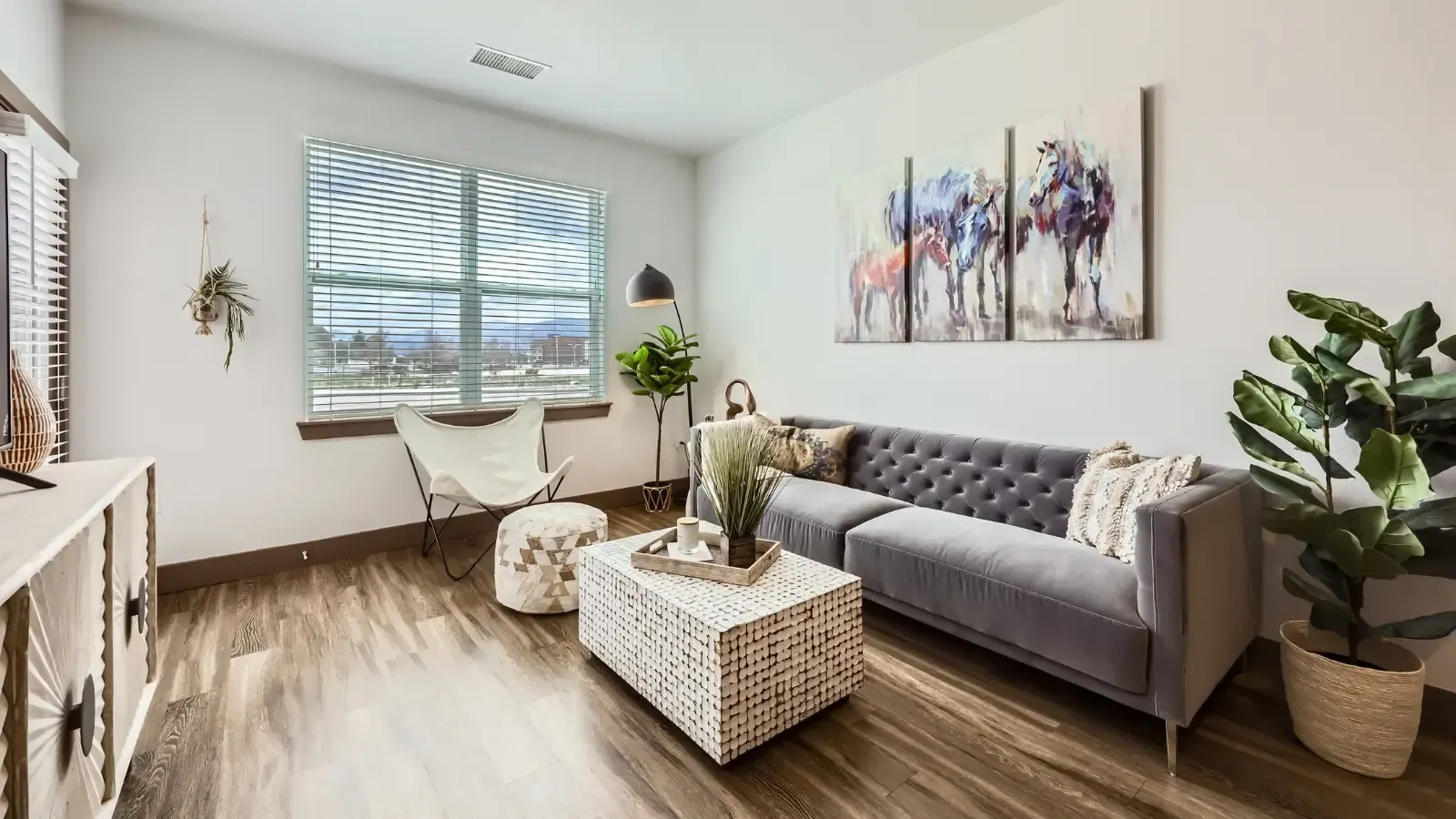 Living room interior of a modern apartment with a gray tufted sofa, white chair, plants, and wall art.