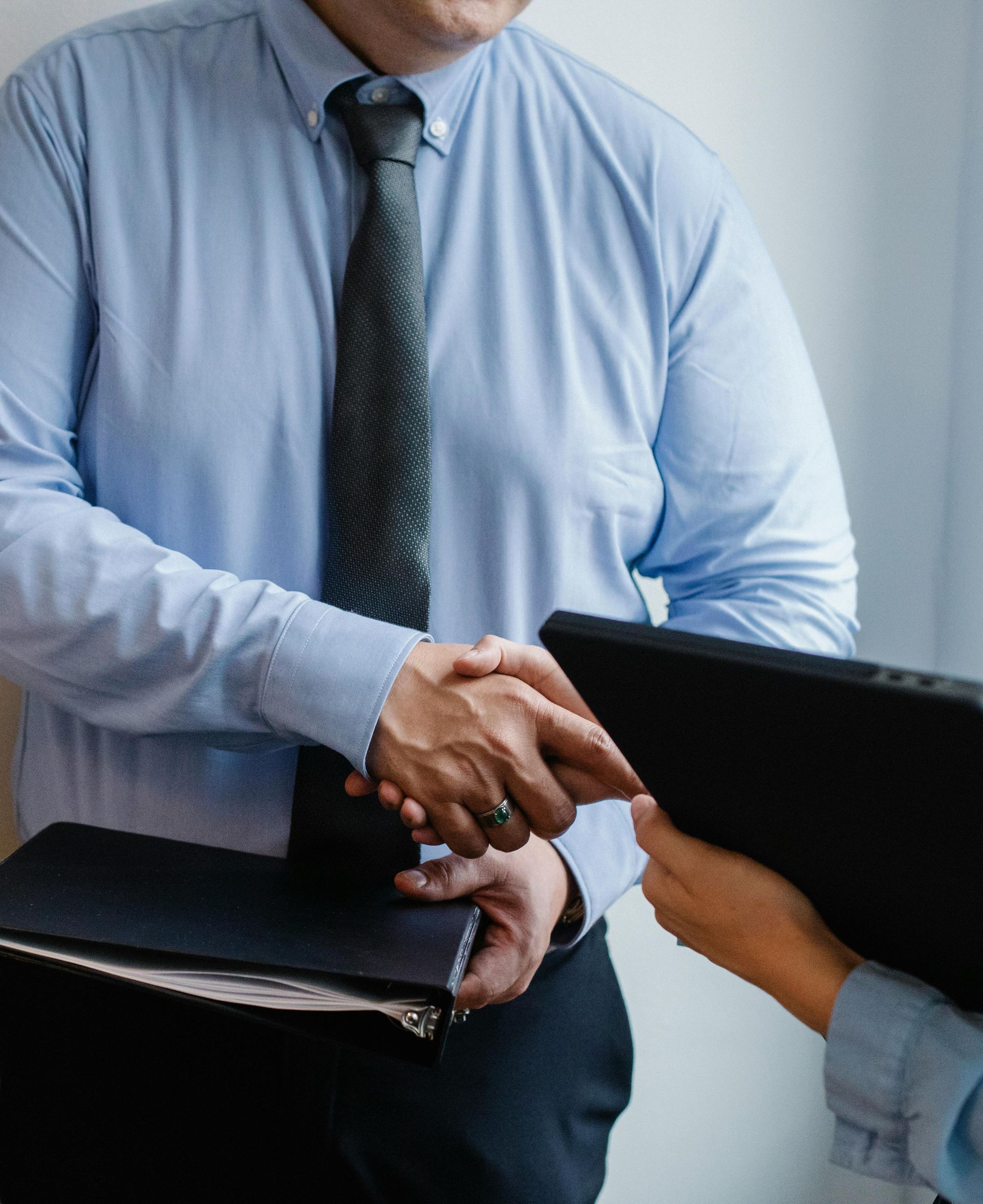 Man in blue shirt and tie shakes hands with another person, holding a folder.
