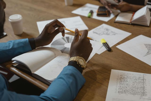 Hands of a person in a blue blazer with a gold watch holding a pencil over an open book at a desk with papers.