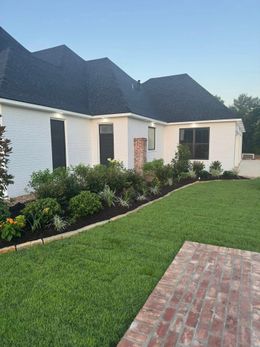 White brick house with dark roof, manicured lawn, and garden bed.