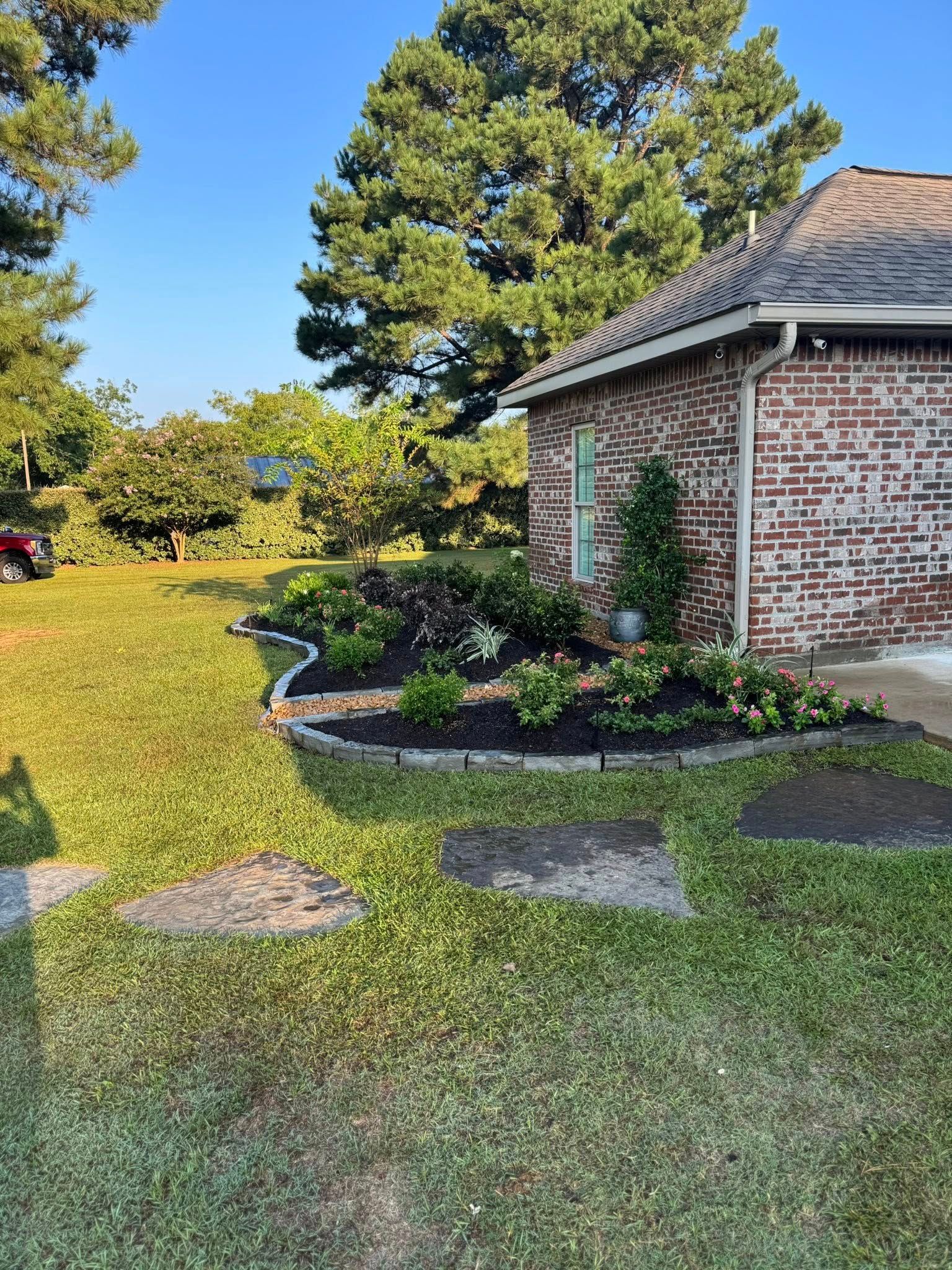 Exterior view of a brick house with a landscaped garden bed along the side. Green grass and trees in the background.