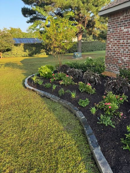 Landscaped flower bed with curved stone edging, next to brick wall and green lawn.
