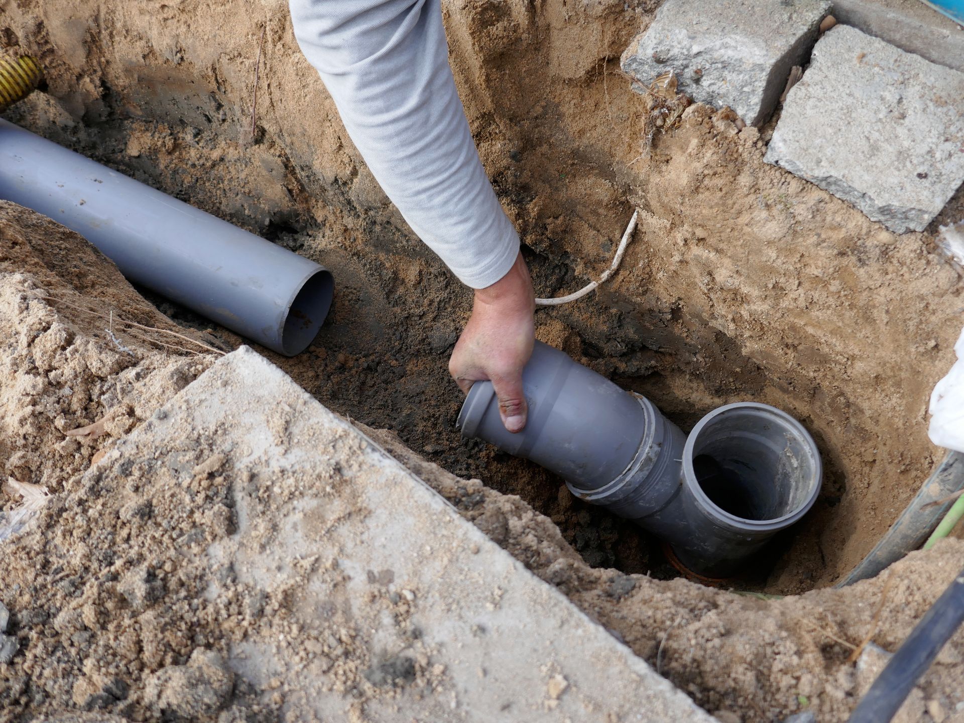Person connecting a gray PVC pipe to a drain fitting in a dirt trench outdoors.