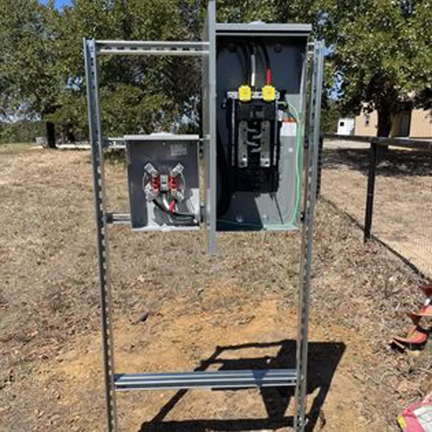 Electrical panel mounted on a metal frame, outdoors. Contains wires and circuit breakers. Gray box, green ground wire.