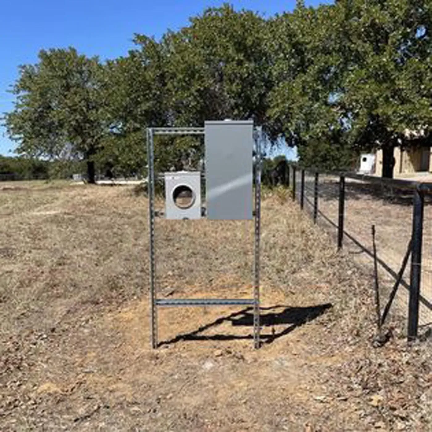 Metal electrical box on a metal frame, in a dry, grassy field with a fence and trees in the background.