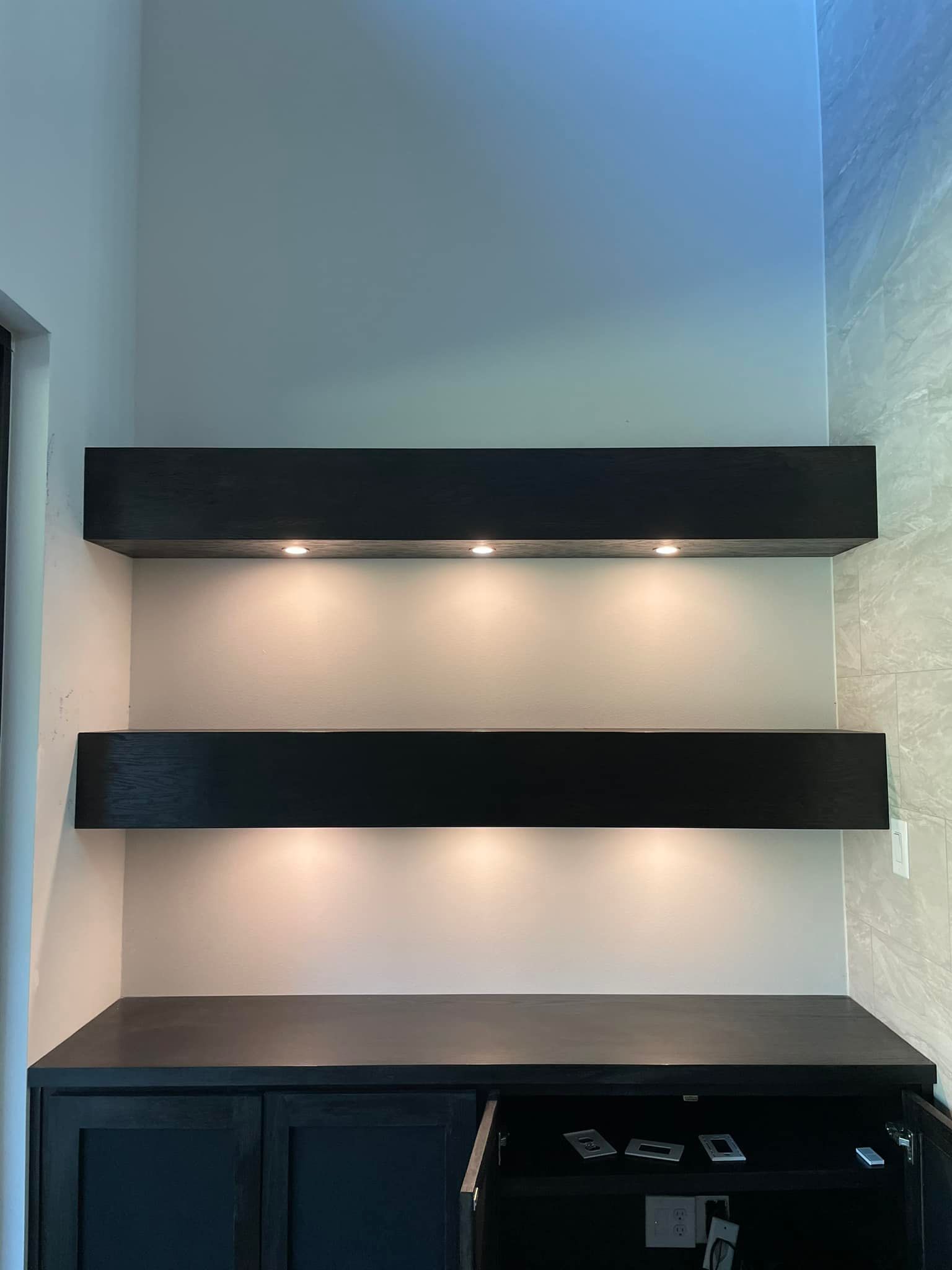 Black floating shelves with inset lights above a dark cabinet in a neutral-colored room.