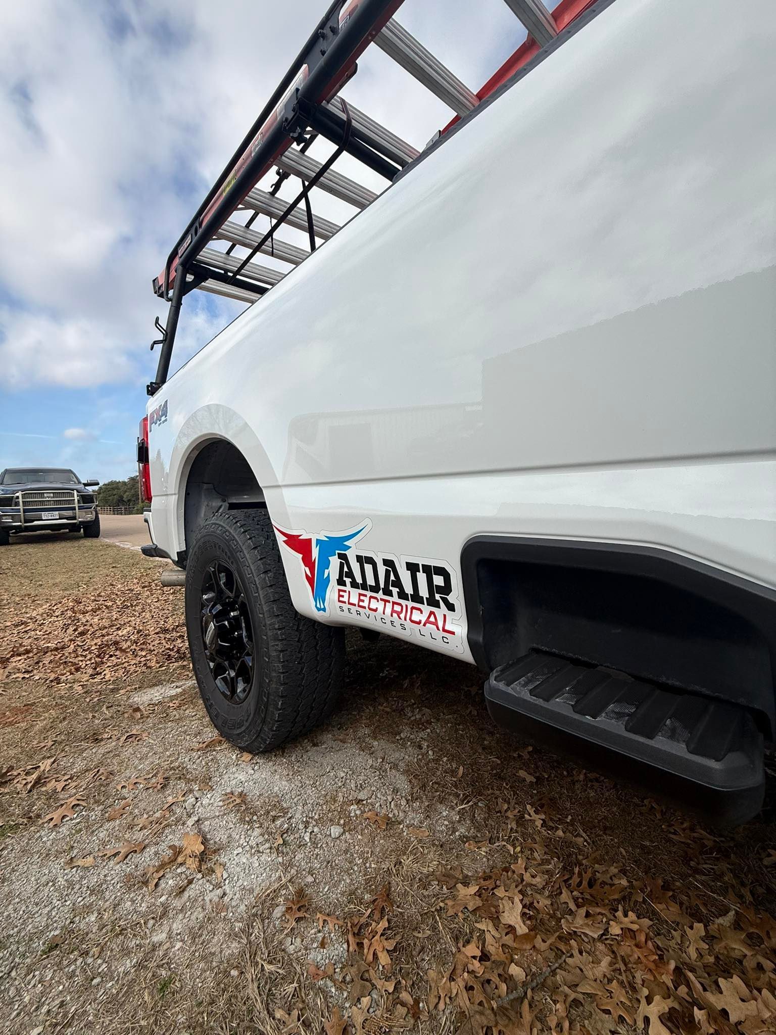 White work truck with ADAIR logo, ladder rack, and black wheels parked outdoors on a cloudy day.