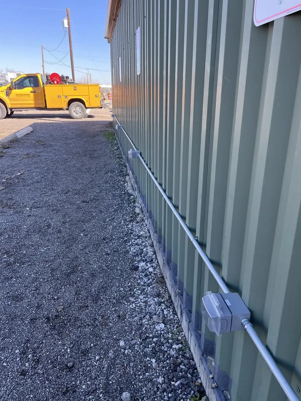 A corrugated metal building with electrical conduit along the wall, gravel path, and yellow truck in the background.