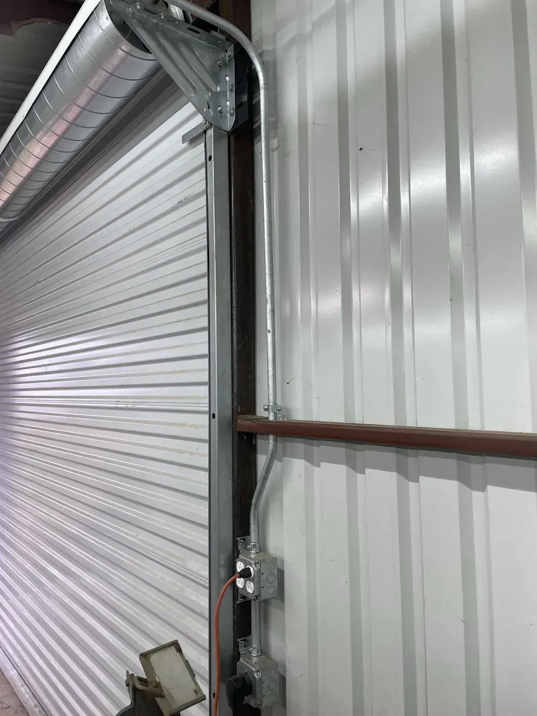 Metal roll-up door inside a corrugated metal-walled building. Electrical conduit and outlet box are visible.